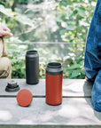 Open red travel tumbler next to an open black travel tumbler resting on bench between two people.