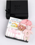 BOXFOX black gift box next to open box containing a book, swaddle, teether, baby beanie and bunny rattle.