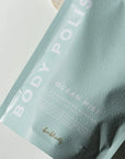 Rectangular, sealed, light blue plastic bag with white text that reads "BODY POLISH" on the side and "OCEAN MIST MOISTURIZING EXFOLIANT" in bottom right. Photographed on grey background.