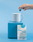 Pink Hibiscus Tea Sachet leaning against blue box with blue mug resting on top.