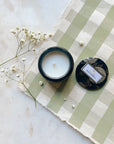 Moso Bamboo glass jar candle with tin lid off to the side, resting on green gingham paper with baby's breath to the side.