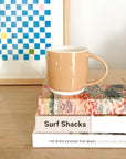 An almond shell bisque ceramic mug sitting on a stack of coffee table books on a light natural wood table in front of a blue and white checkered framed art print.
