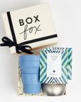 Creme BOXFOX gift box with a bow, next to an open gift box containing a blue collapsible cup, a silver tin candle and two art of tea sachets.