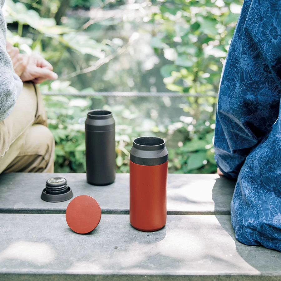Open red travel tumbler next to an open black travel tumbler resting on bench between two people.