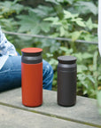 Red Travel Tumbler 500ml next to a black one, sitting on a bench between two people.