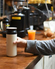 Hand holds white travel tumbler at coffee shop.