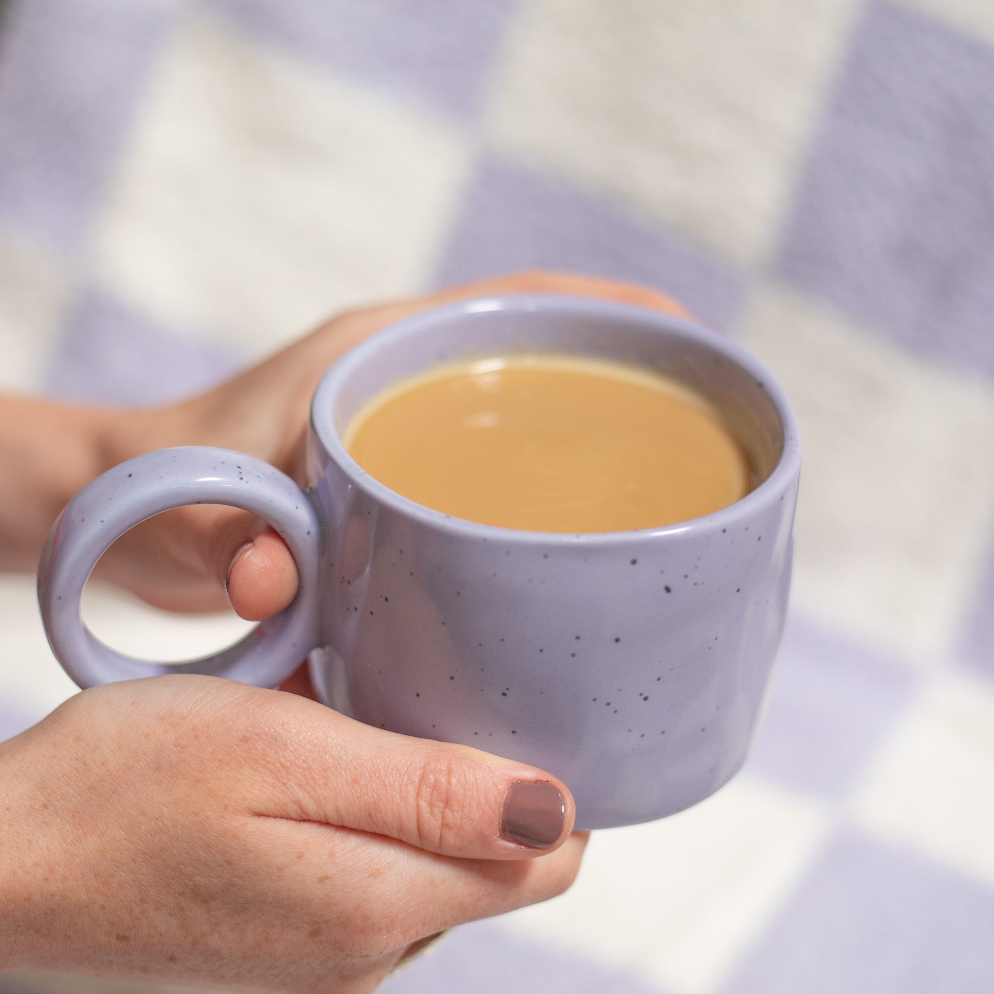 A woman's hands holding a Purple Speckled Nordic Mug over a purple and white checkered plush blanket.