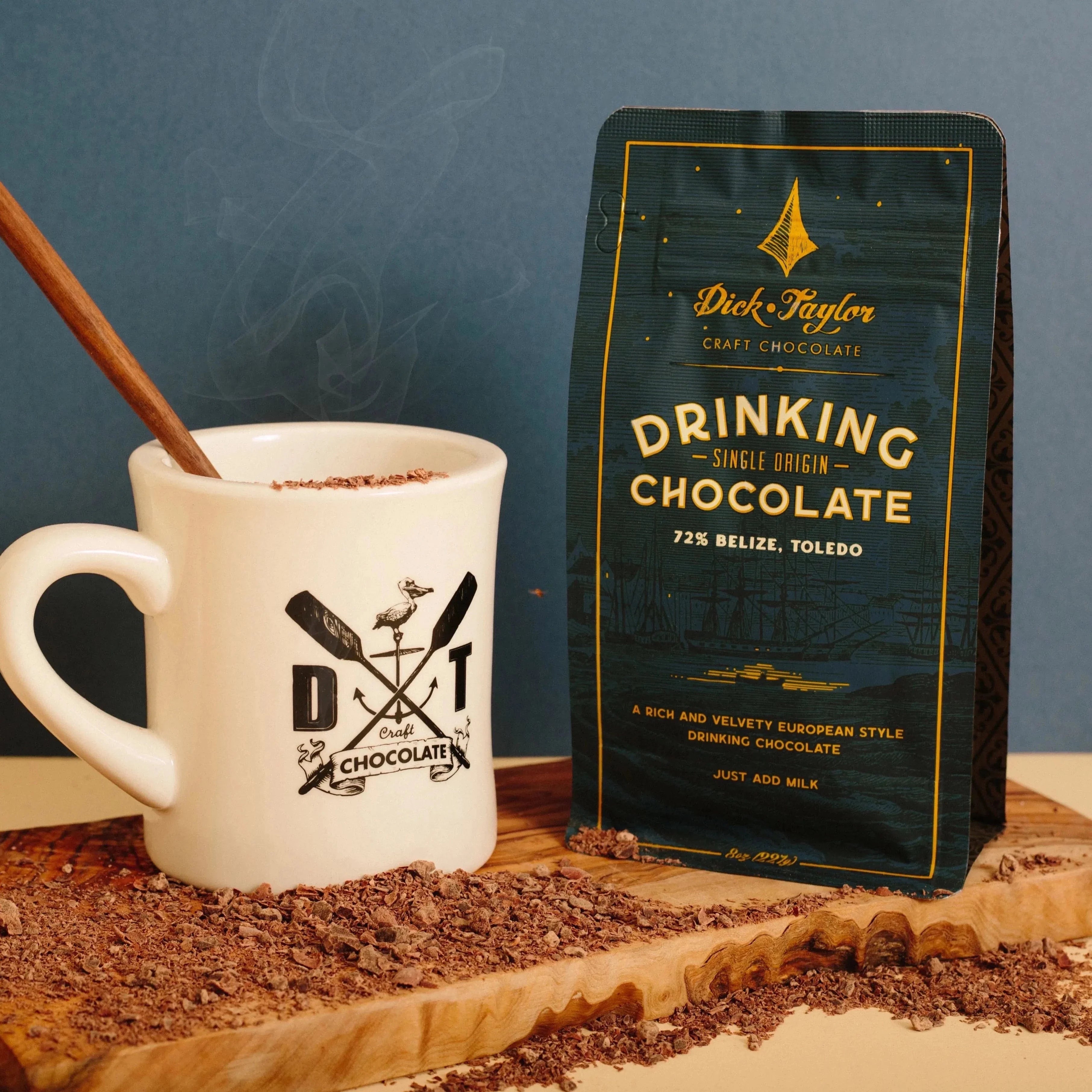 white mug with wooden spoon in it full of drinking hot chocolate. Blue bag of drinking chocolate to the right of it. Both items are on wooden cutting board with a blue background 