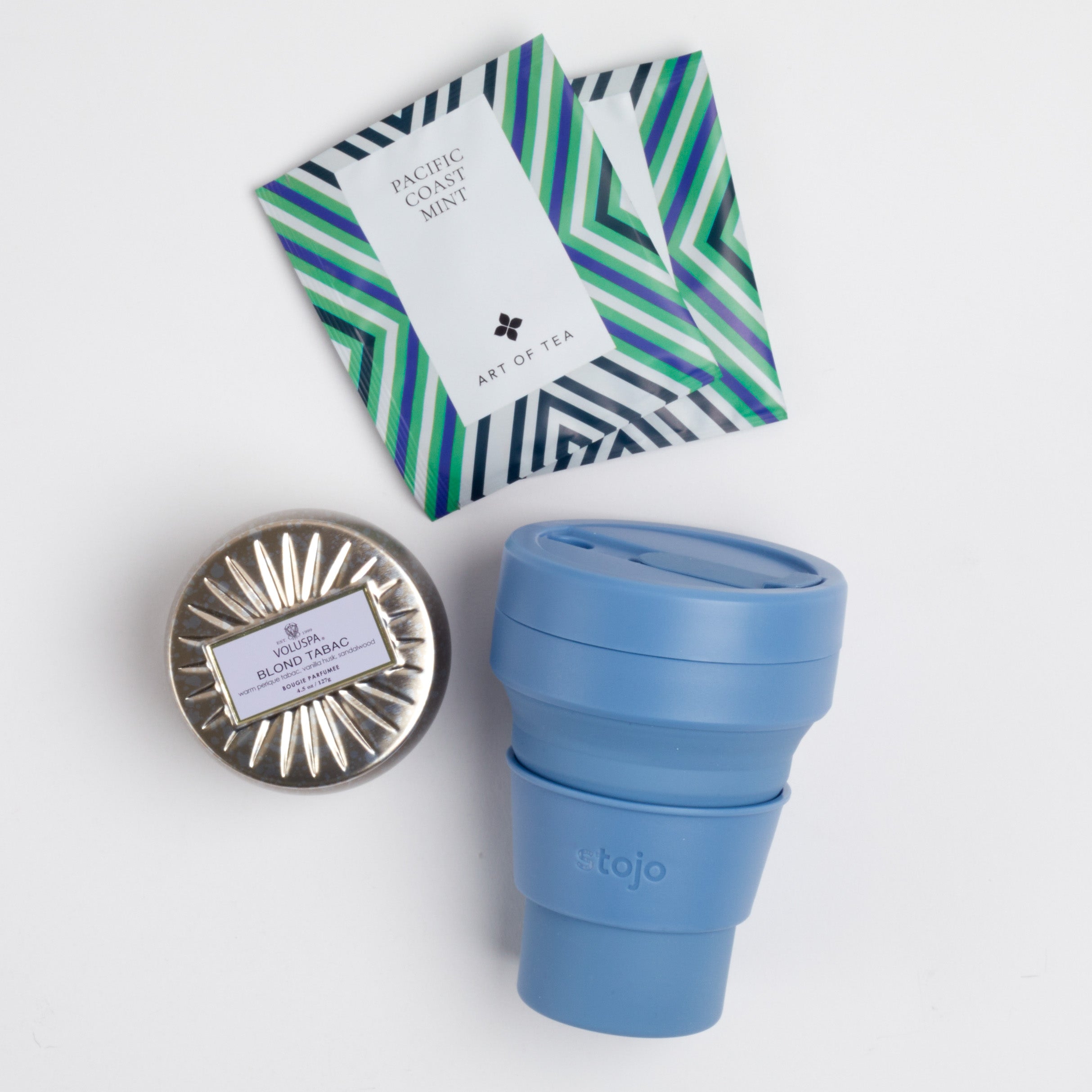 Blue collapsible cup, two art of tea sachets and silver tin candle on white background.