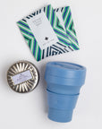 Blue collapsible cup, two art of tea sachets and silver tin candle on white background.