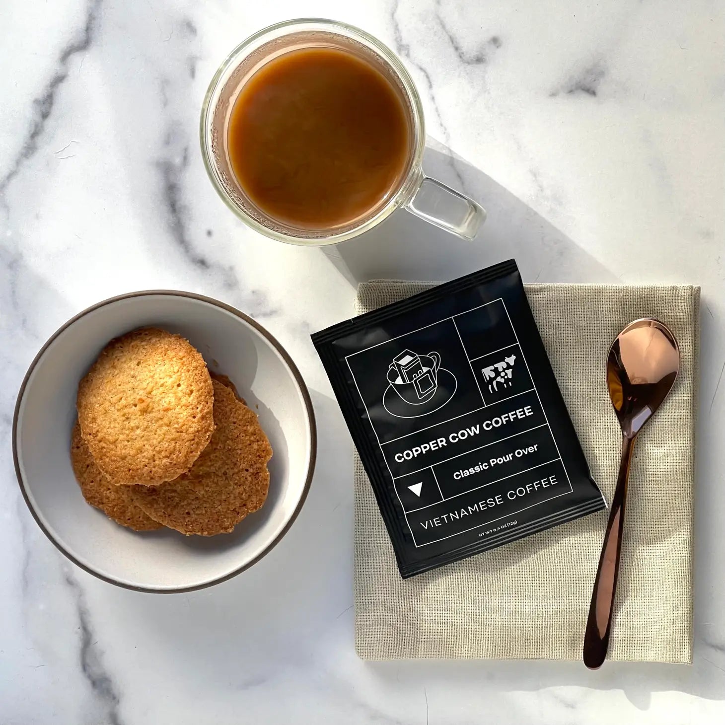 Black packaging for single serving coffee next to a spoon, plate of cookies and mug of coffee.