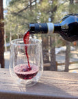 Clear wine glass sits on wooden railing as red wine is poured into it. This overlooks pine trees.