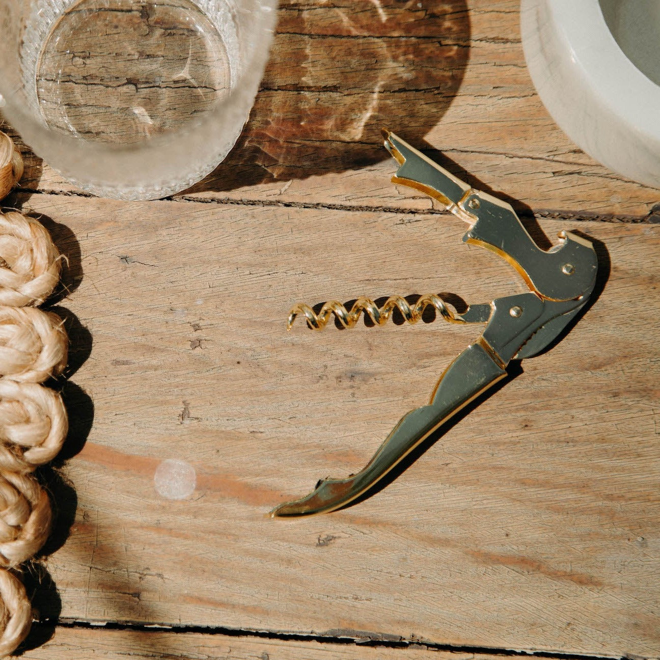 A gold Mielle wine opener laying on a wooden table next to a clear glass of water.