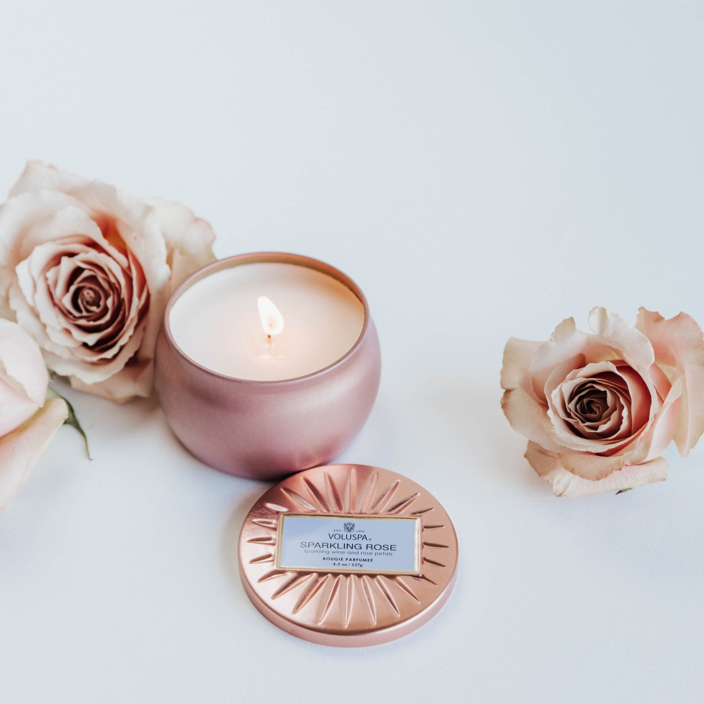 Pink sparking rose tin candle lit next to dusty pink roses.