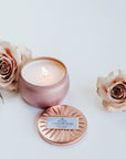 Pink sparking rose tin candle lit next to dusty pink roses.