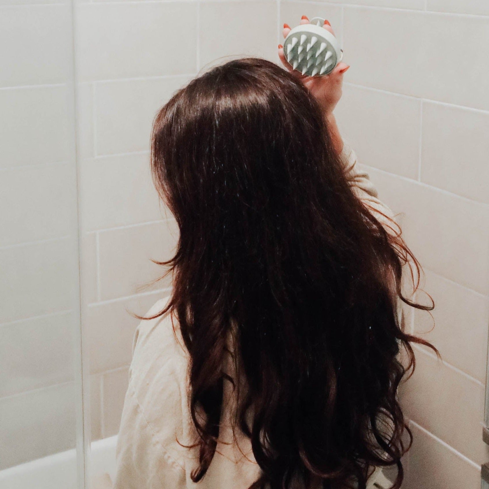 A woman with long wavy brown hair using a sage green scalp massager to massage the back of her head.