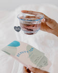 Hand hold blue glass tea cup with beauty tea sachet next to beauty tea packaging.