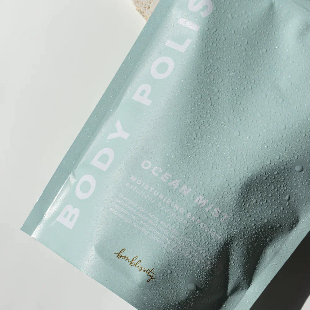 Rectangular, sealed, light blue plastic bag with white text that reads &quot;BODY POLISH&quot; on the side and &quot;OCEAN MIST MOISTURIZING EXFOLIANT&quot; in bottom right. Photographed on grey background.