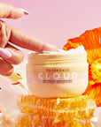 Barrier Repair Cloud Cream surrounded by honeycomb and flowers, with hand sticking finger in tub.