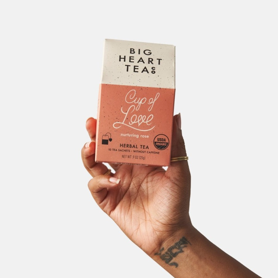 House-shaped cardboard box tea packaging, held by a hand with a ring and tattoo on the wrist. 
