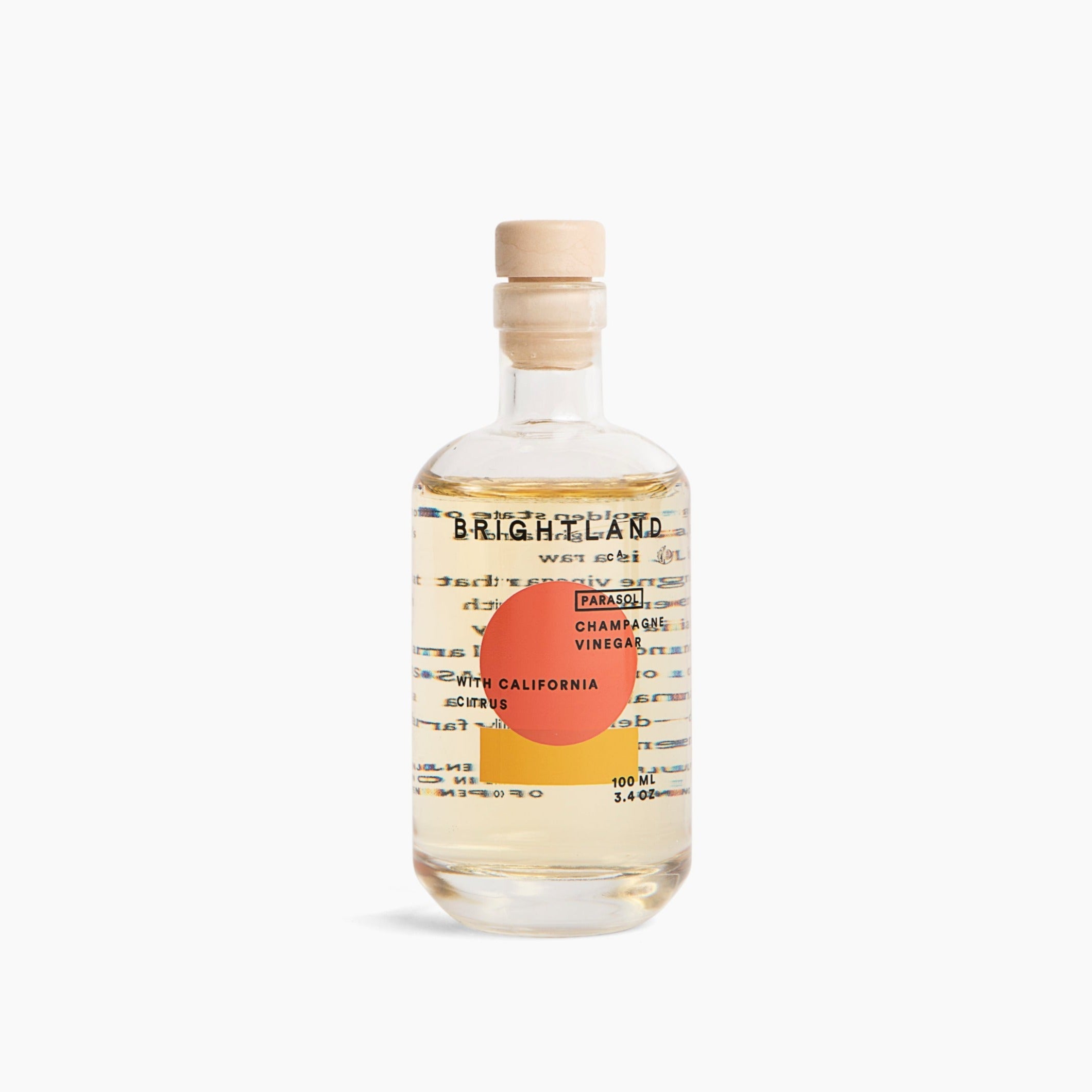 A clear glass bottle with orange and yellow geometric motifs with the following in black writing &quot;BRIGHTLAND CA PARASOL CHAMPAGNE VINEGAR WITH CALIFORNIA CITRUS 100ML 3.4 OZ&quot; photographed on white background.