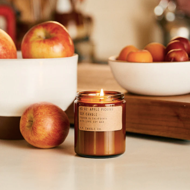 The P.F. Candle Co Apple Picking Candle with lit flame on beige countertop with white bowls of apple behind.