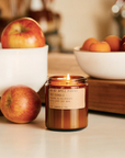 The P.F. Candle Co Apple Picking Candle with lit flame on beige countertop with white bowls of apple behind.