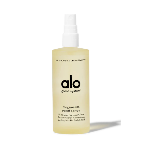 A semi-opaque light yellow plastic spray bottle with white spray nozzle. Black text on bottle reads, &quot;amla-powered clean beauty alo glow systems magnesium reset spray restorative magnesium, amla, arnica &amp; valerian aromatherapy soothing mist for body &amp; mind&quot;. Photographed on white background.