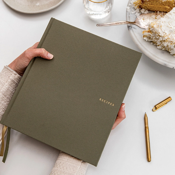 A Caucasian woman&#39;s hands hold olive green linen journal with &quot;RECIPES&quot; embossed in gold on center right over white table that also holds uncapped gold pen, marble platter with cake and silver serving knife and glass cup. 