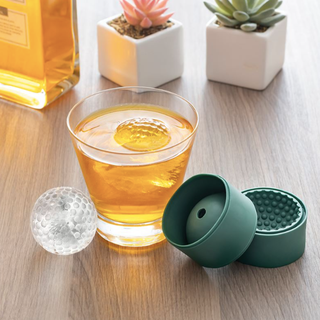 A short drinking glass filled with liquor and holding one pice of golf ball shaped ice sitting on wooden table. Other items on table include: green golf ball ice mold, golf ball shaped ice, two succulents in white ceramic planters, and bottle of liquor. 