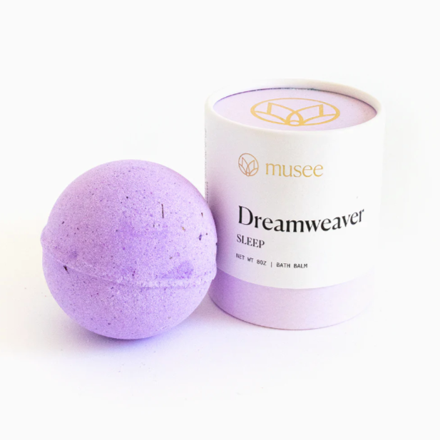 A purple spherical bath bomb to the left of white and purple cylindrical packaging that reads "musee Dreamweaver Sleep Bath Balm" photographed on white background.
