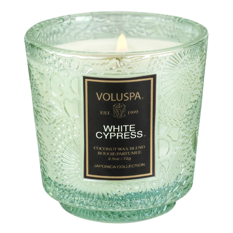 A white candle with single burning wick at top in floral patterned green glass holder. Black label in center reads, &quot;Voluspa Est. 1999 White Cypress Coconut Wax Blend Bougie Parfumee 2.5oz/72g Japonica Collection&quot;.