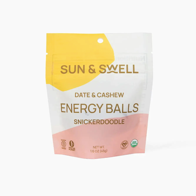 A white resealable plastic bag with yellow and pink oblong shapes on opposite corners with brown text that reads, &quot;Sun &amp; Swell Date &amp; Cashew Energy Balls Snickerdoodle&quot; photographed on white background.