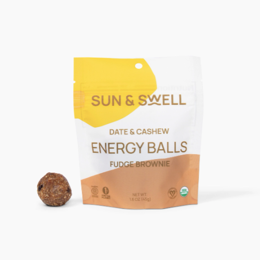 A white resealable bag with yellow and brown oblong shames on opposite corners of bag. Brown writing on bag reads, &quot;Sun &amp; Swell Date &amp; Cashew Energy Balls Fudge Brownie&quot;. Photographed on white background.