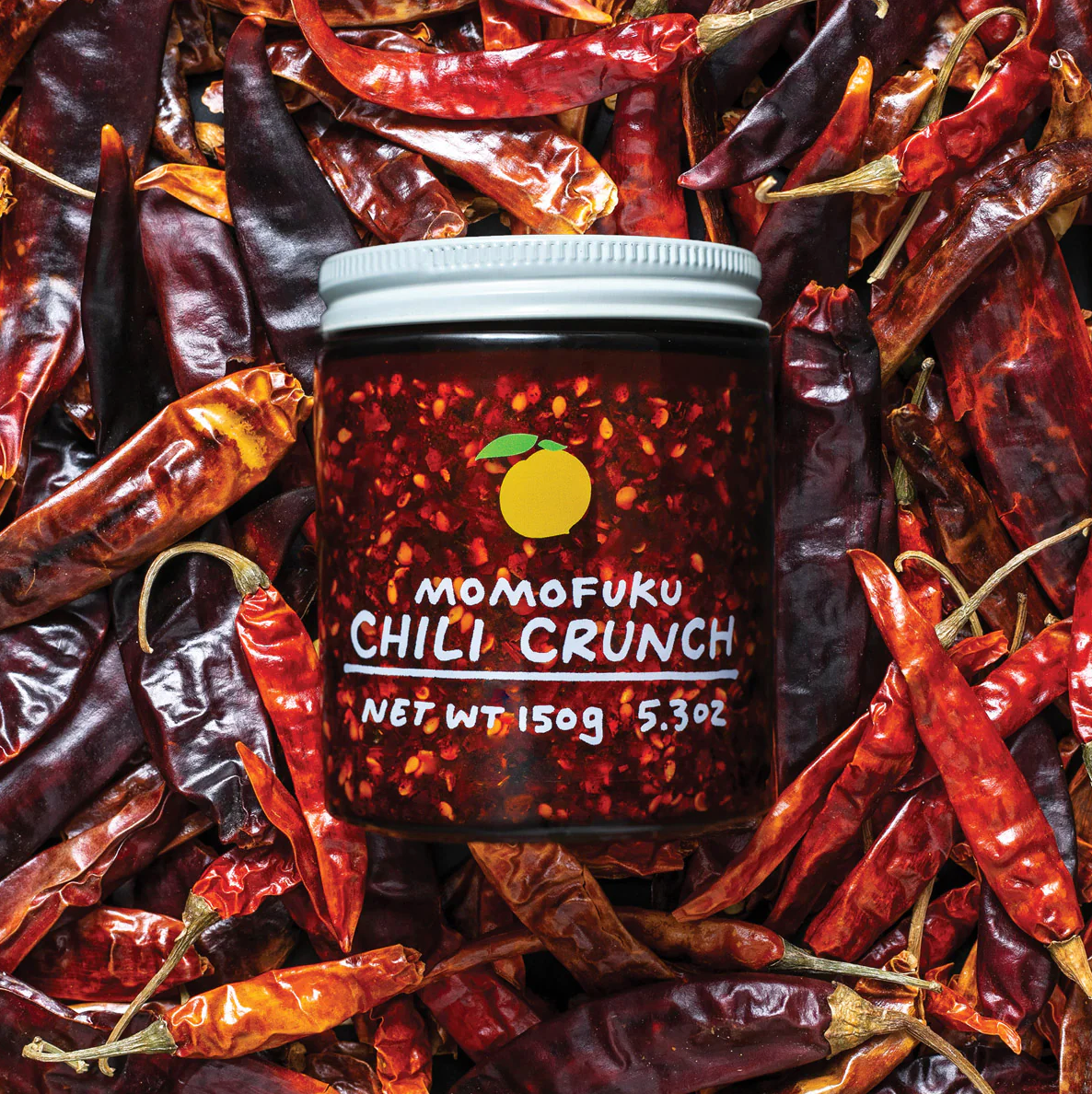 Image shows Momofuku&#39;s Chili Crunch in a clear jar with white lid, lying on a bed of chiles. 