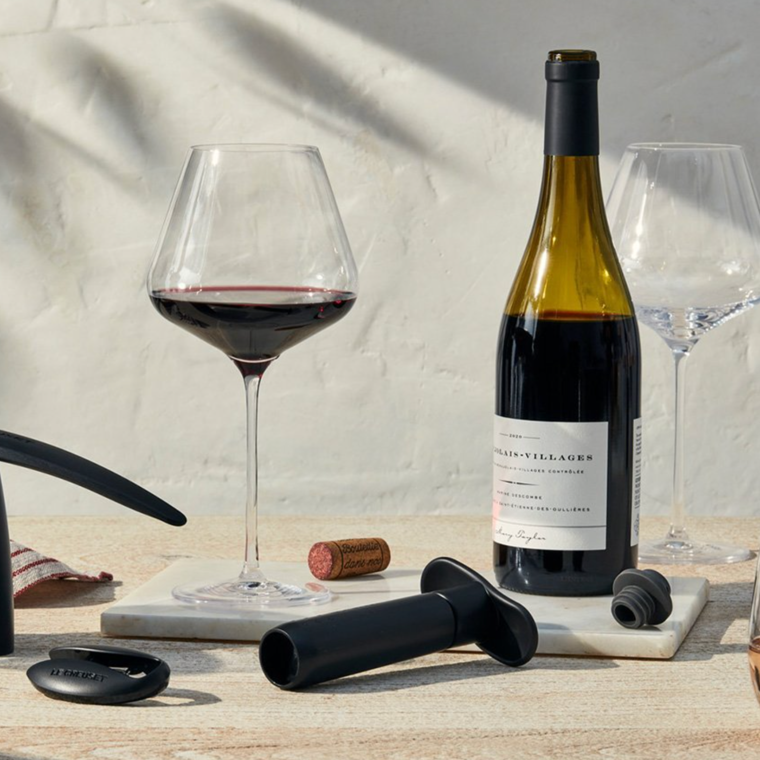 Metal Wine Pump and 3 Stoppers next to bottle of red with glasses and corks.