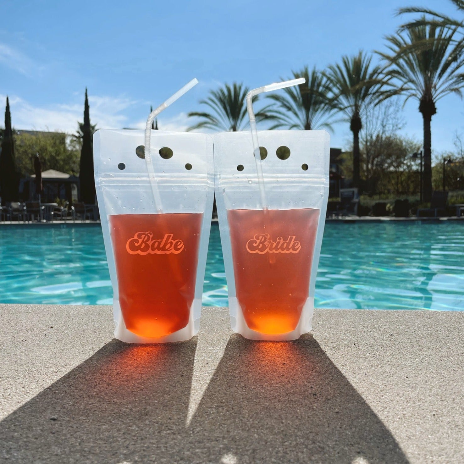Two transparent plastic drink pouches standing upright next to a pool of water. Each pouch is holding a pink colored liquid. Water and palm trees are in the background as the pouches sit on a cement poolside. The left pouch says &quot;Babe&quot; in pink cursive text and the right pouch says &quot;Bride&quot; in white cursive text.