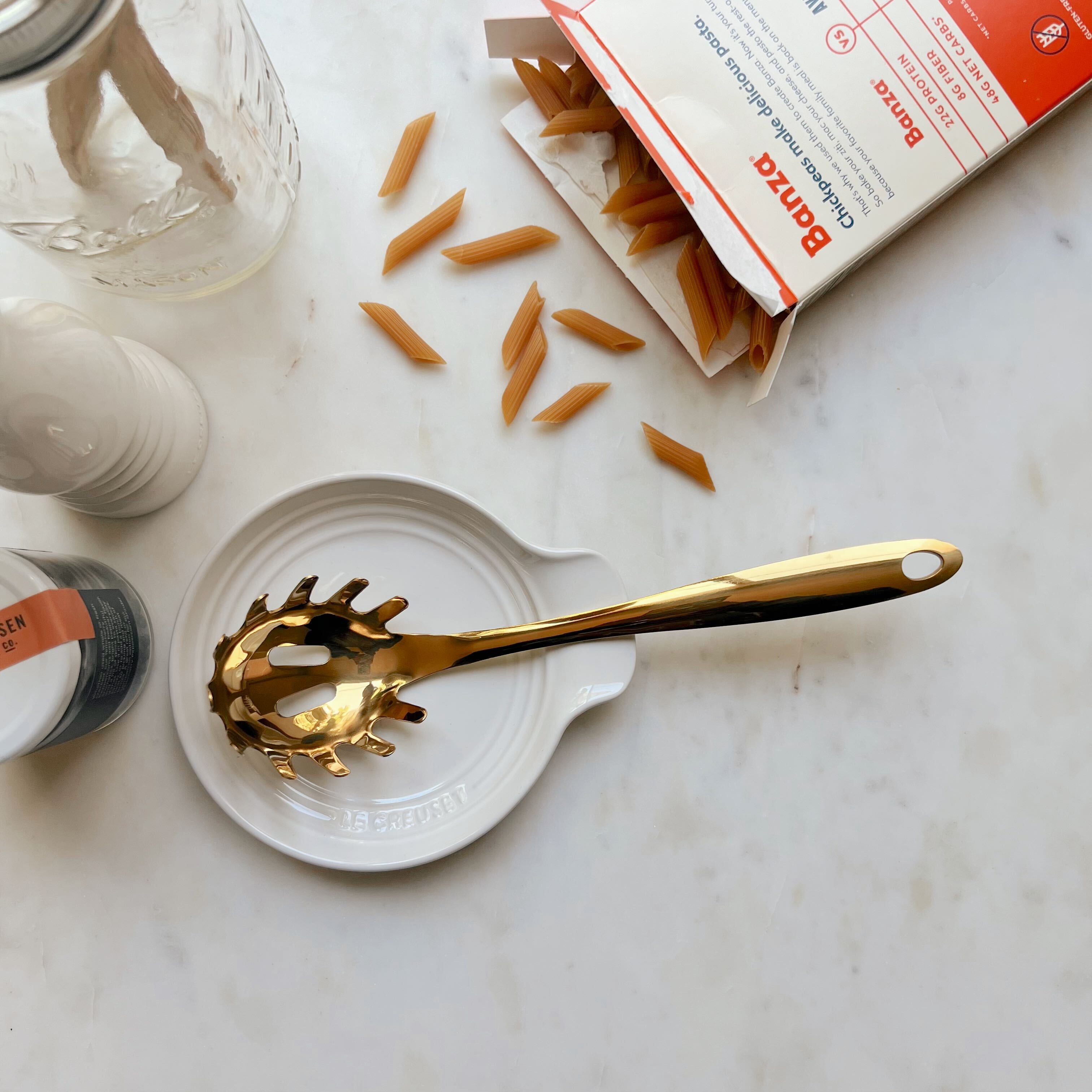 A gold pasta spoon laying on a white ceramic Le Creuset spoon rest next to Banza pasta noodles spilling out of their package on a white marble counter.