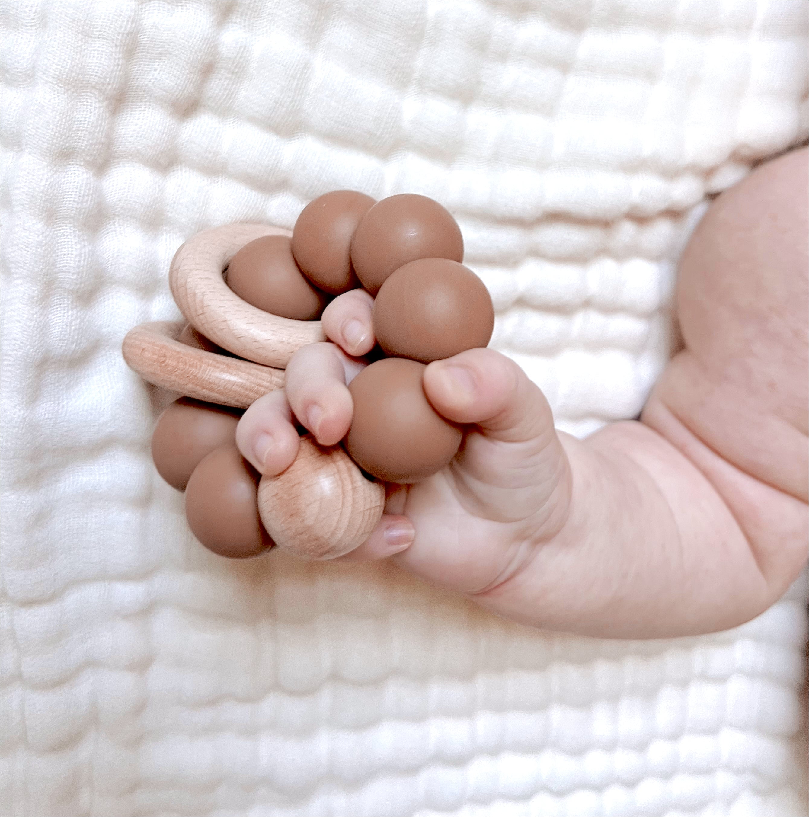 a baby holding a terracotta colored silicone ring teether laying on a white muslin blanket