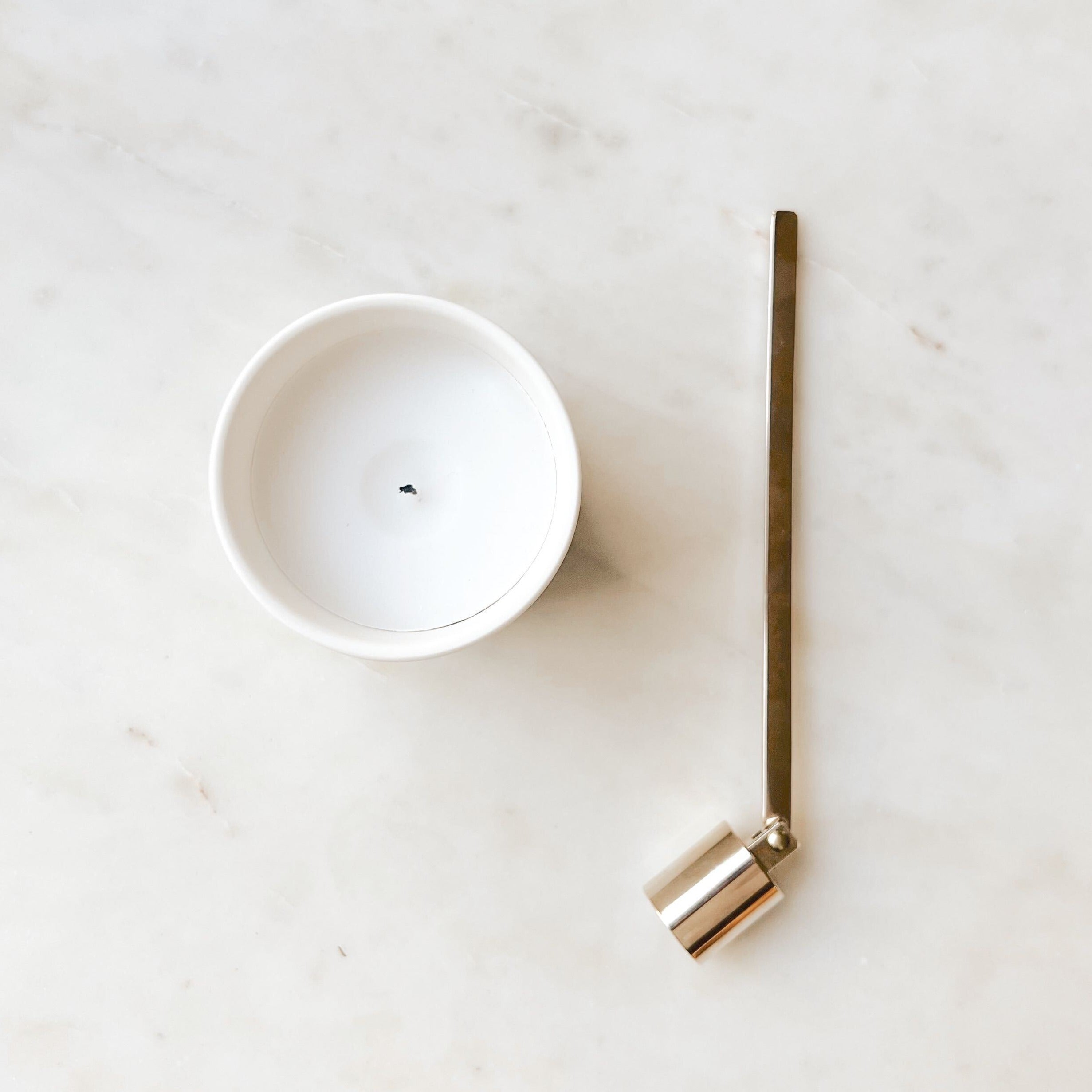 A gold candle snuffer laying next to a white Fiat Lux Copenhagen candle on a white marble table.