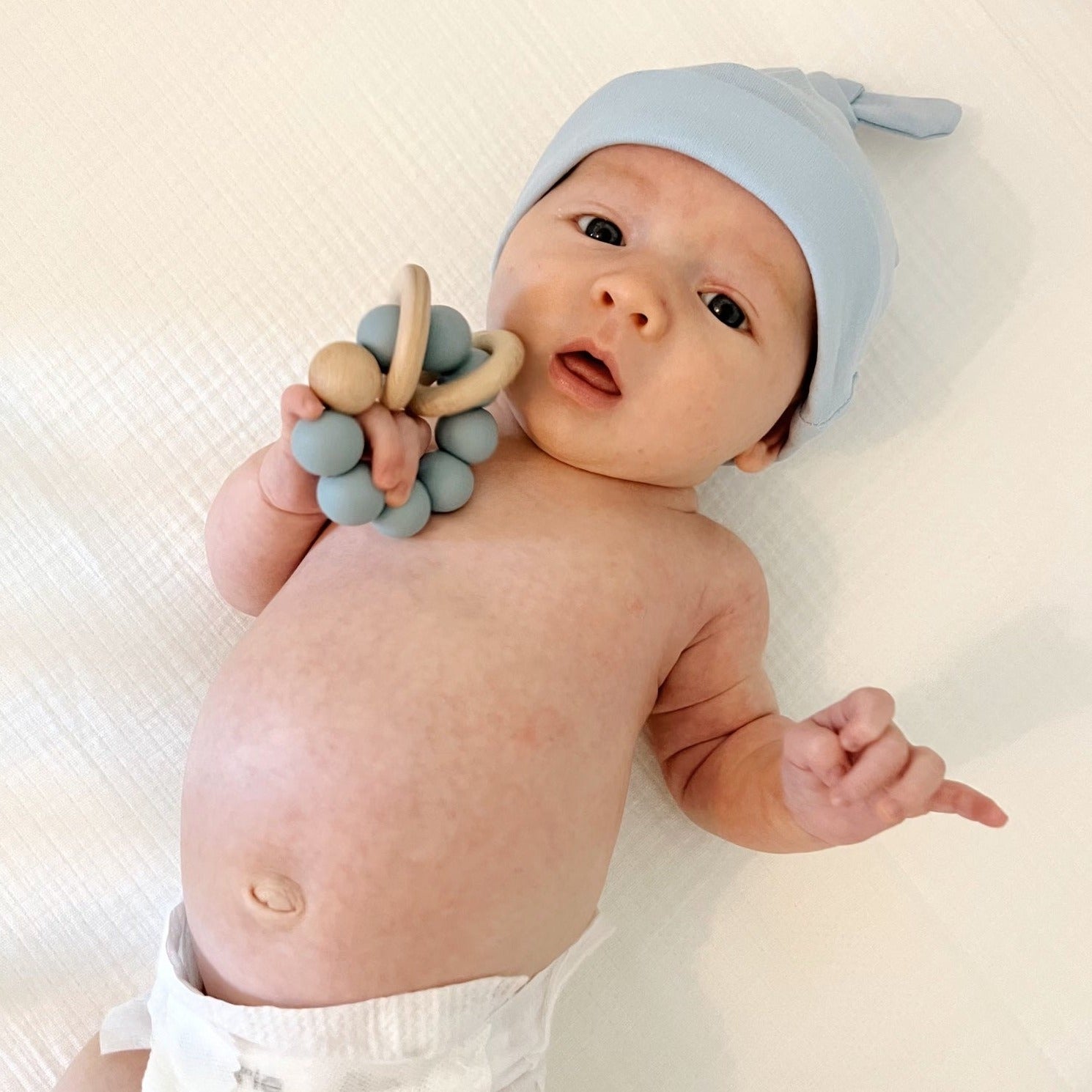 A baby wearing a blue topknot cotton hat and holding a blue silicone baby teether while laying on a white muslin crib sheet.