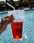A woman's hand holding a transparent drink pouch holding pink colored liquid in front of a pool of water. On the pouch is cursive lettering spelling out the word "Babe" in light pink text.