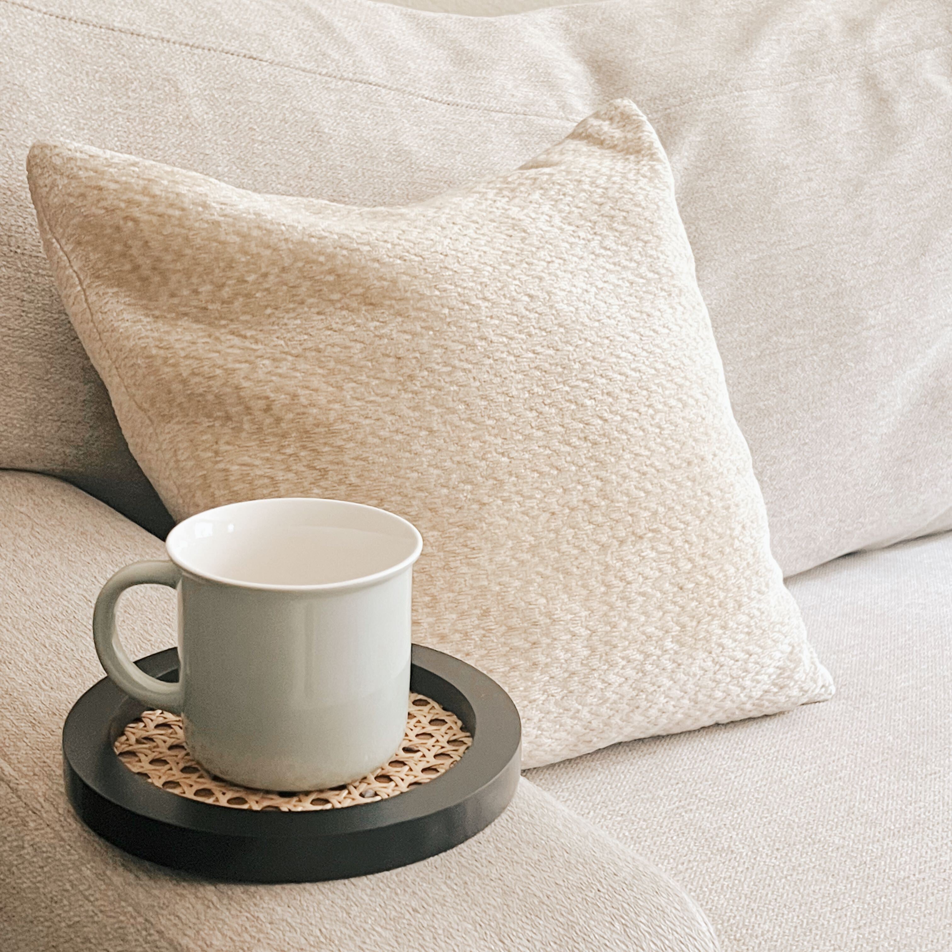 A black rattan cane tray holding a grey ceramic mug on the armrest of an oatmeal colored couch.