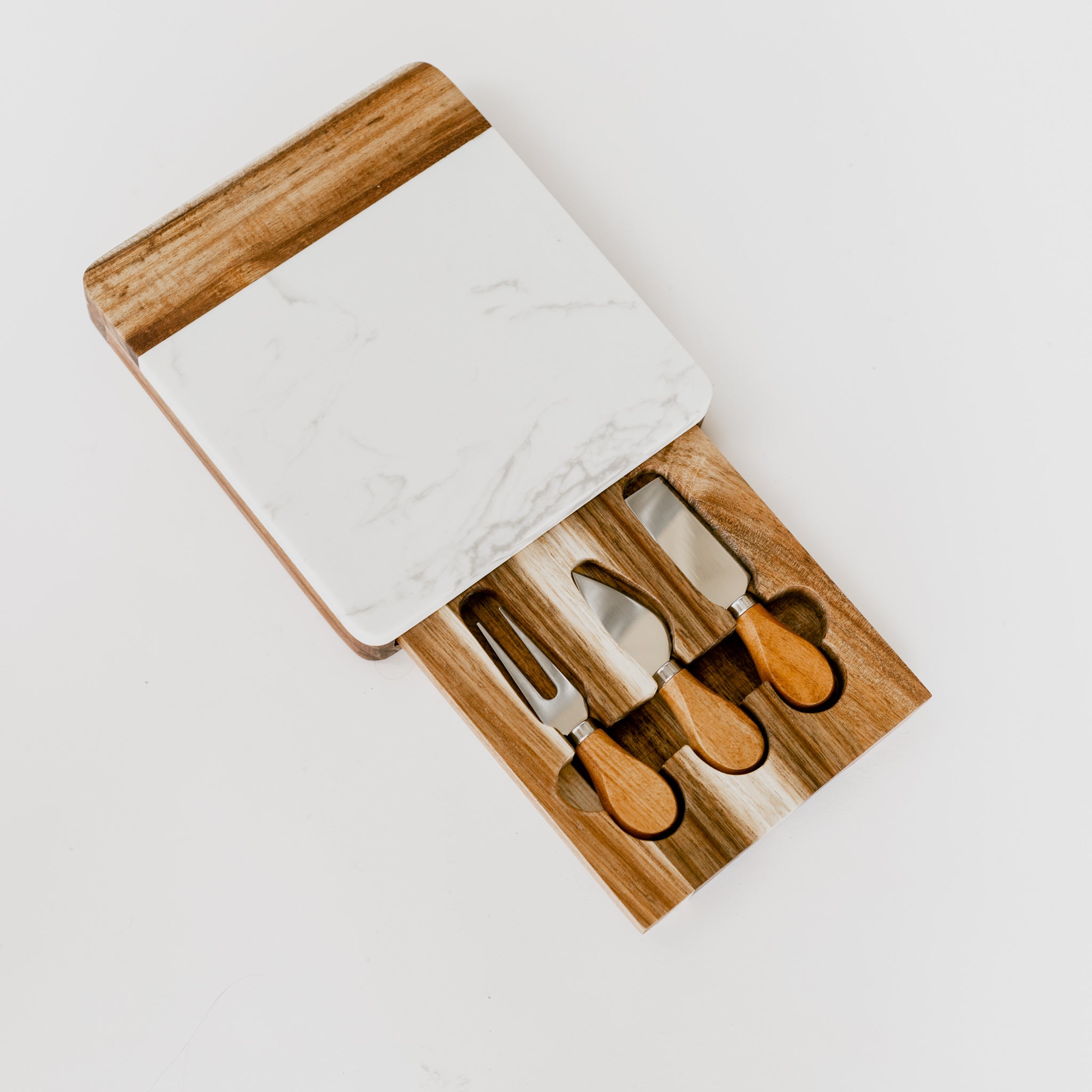 A white marble and acacia wood charcuterie board with a pull out drawer holding 3 stainless steel cheese knives against a white background
