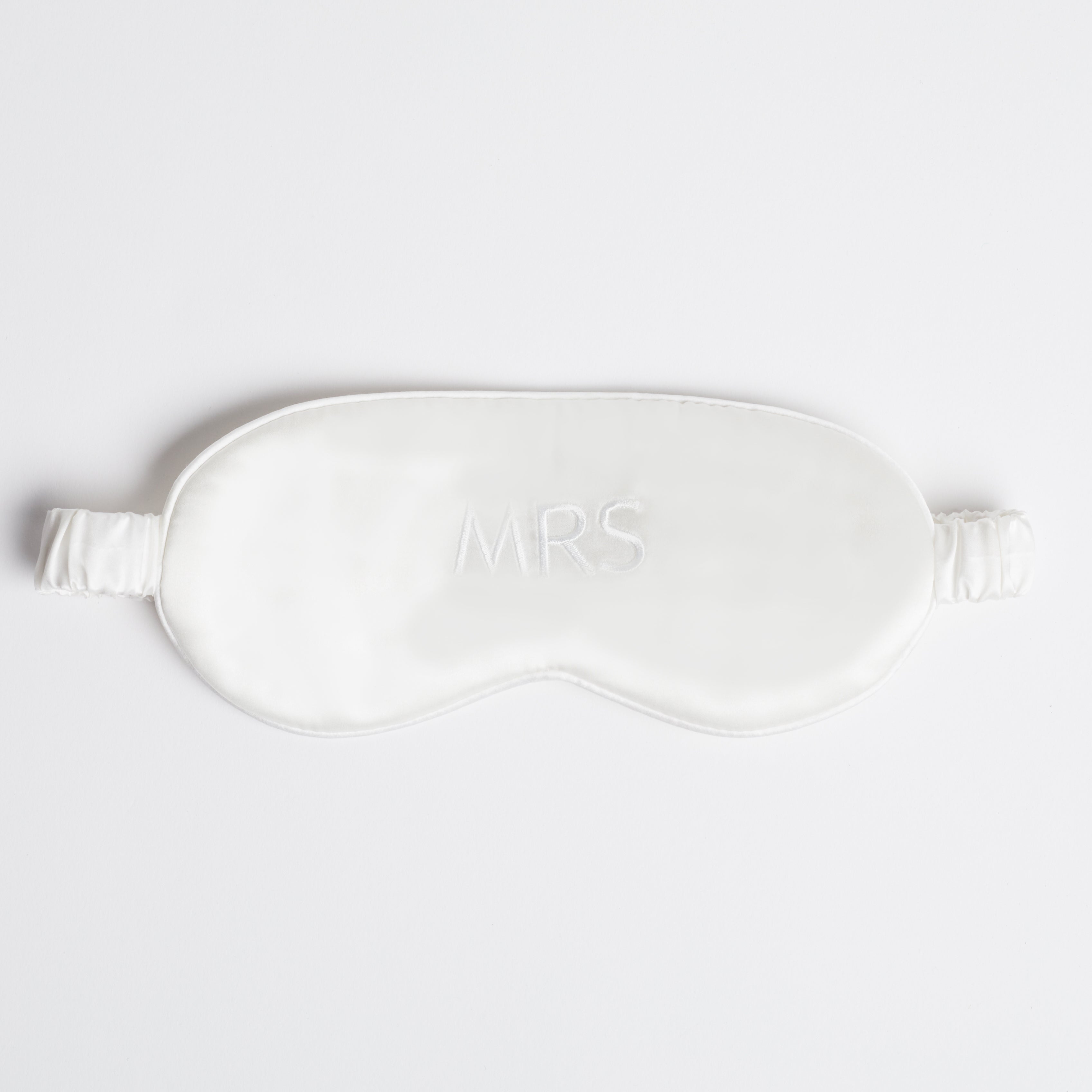 White silk eye mask with "MRS" written across it, lying on a white background.