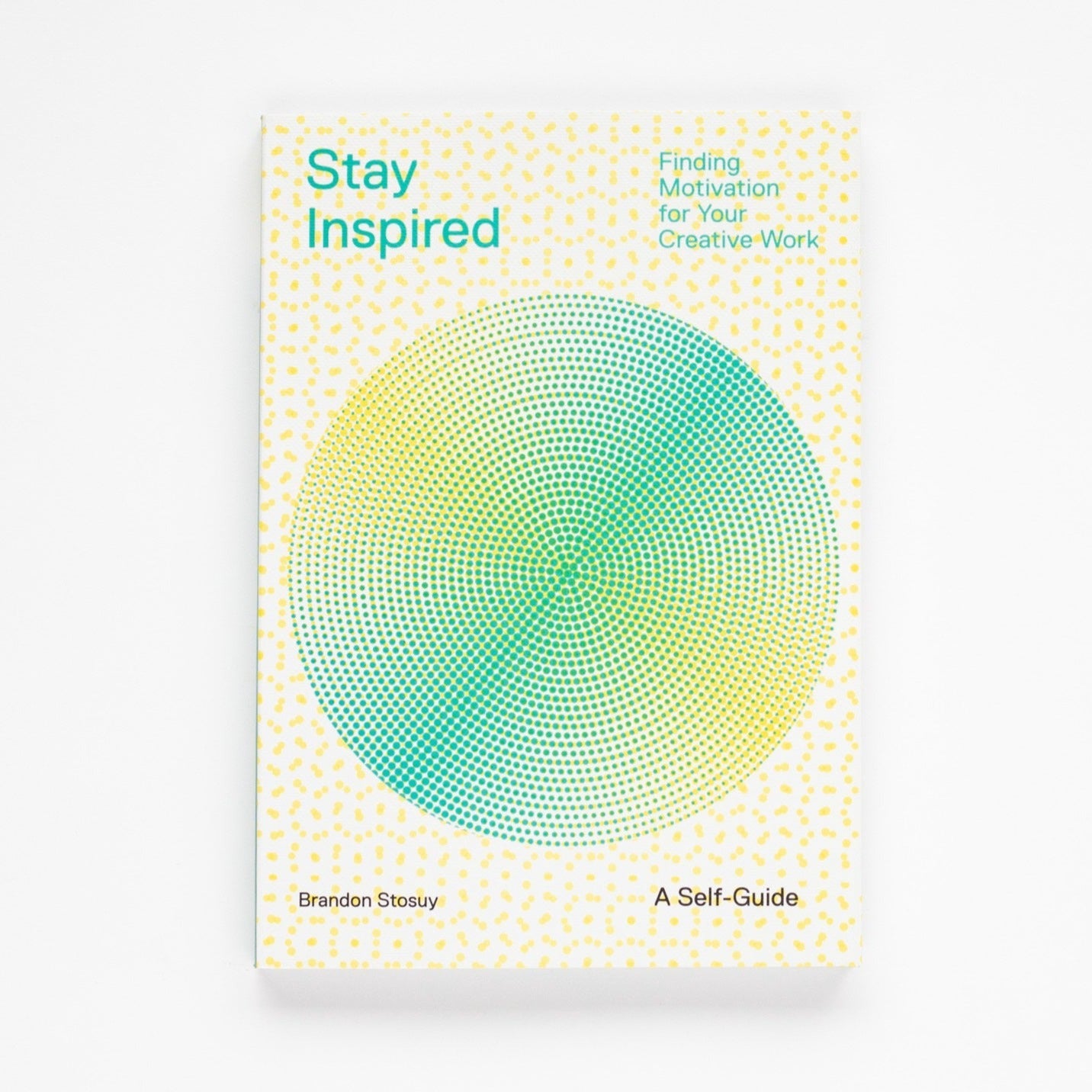 A cream colored paperback book with yellow geometric design all over and yellow and green circle in middle. Green and black text on book reads, "Stay Inspired Finding Motivation for Your Creative Work Brandon Stosuy A Self-Guide". Photographed on white background.