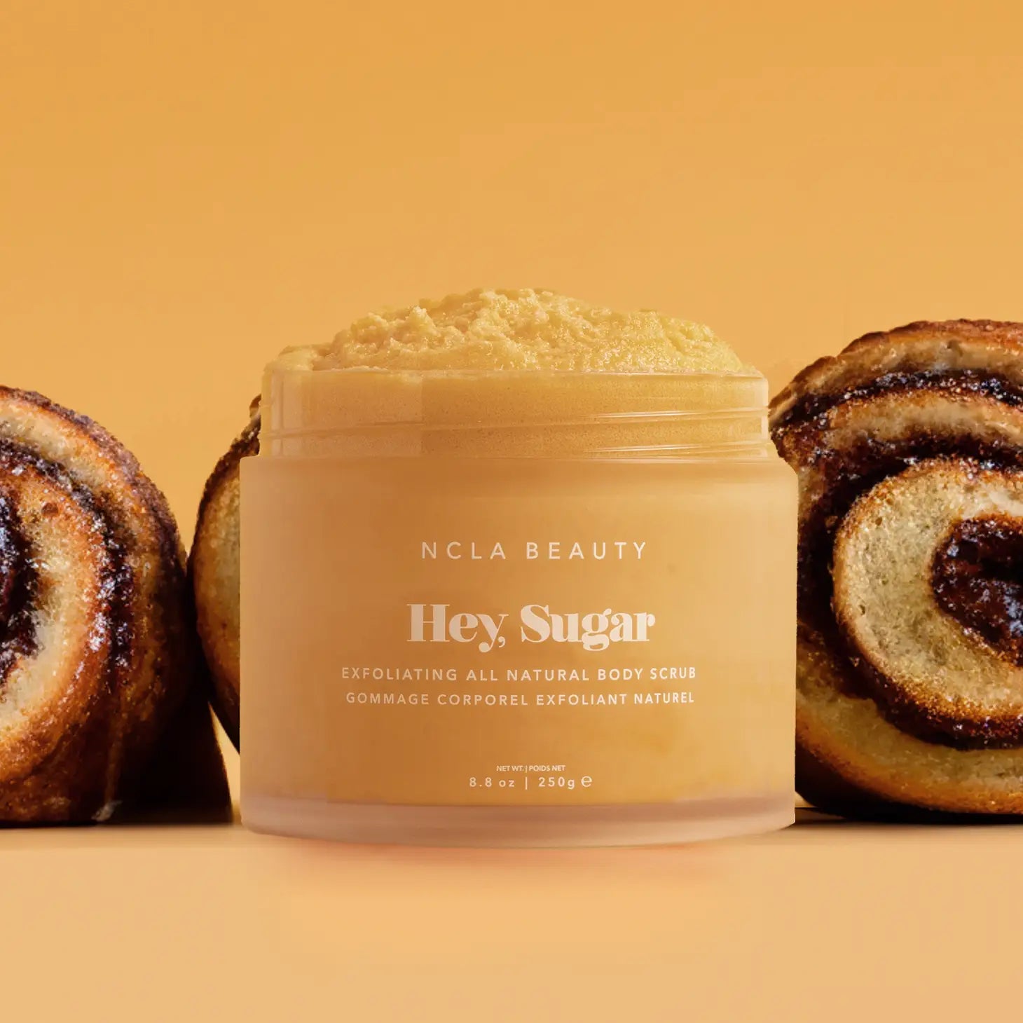 Cinnamon Roll Body Scrub sits in front of row of cinnamon rolls on brown background.