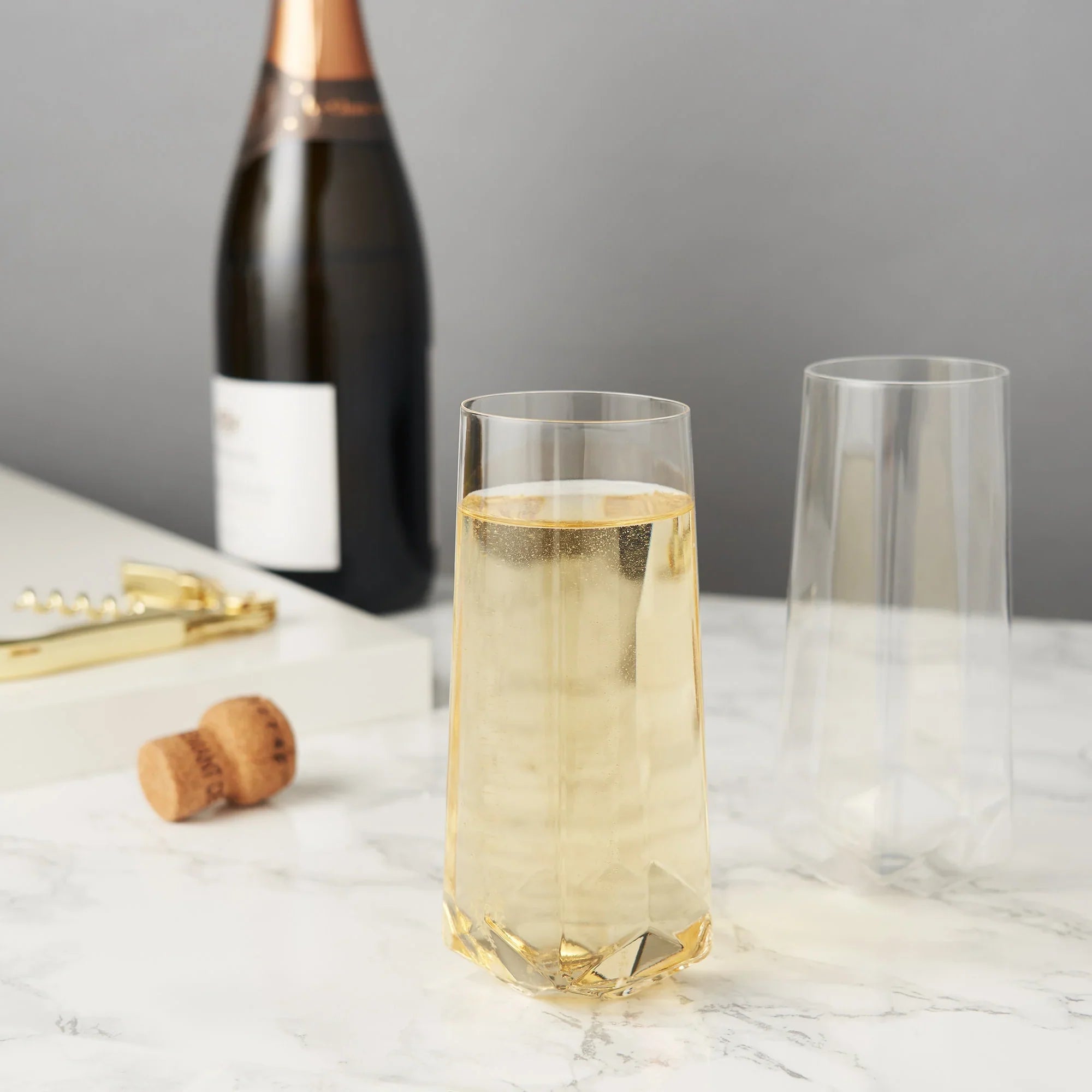Faceted Crystal Stemless Champagne Flutes filled with champagne, with champagne bottle, cork and bottle opener in the background