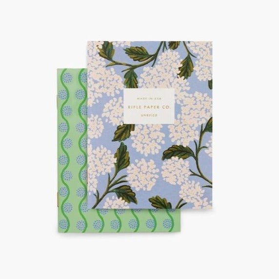 Small notebook with periwinkle background and pink and white hydrangea flowers. The second notebook is green with blue wavy lines and small dots.