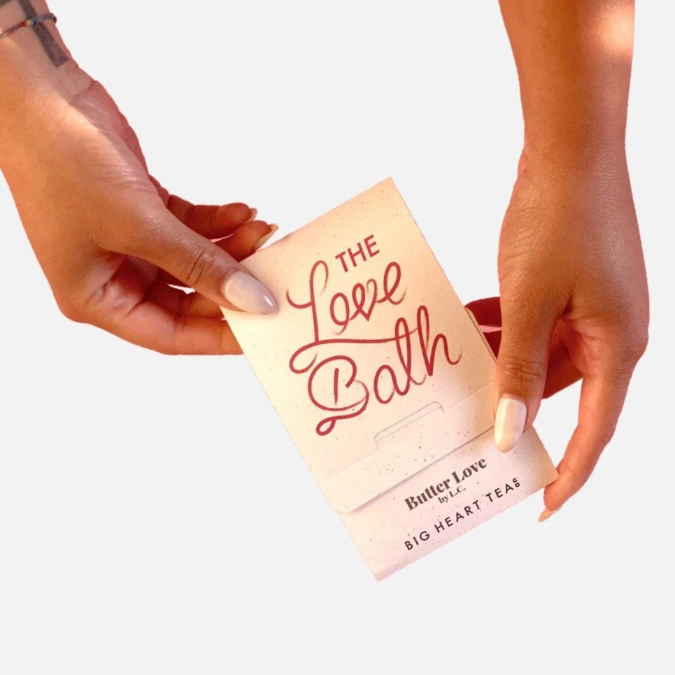 Two hands holding white The Love Bath packaging featuring red font.